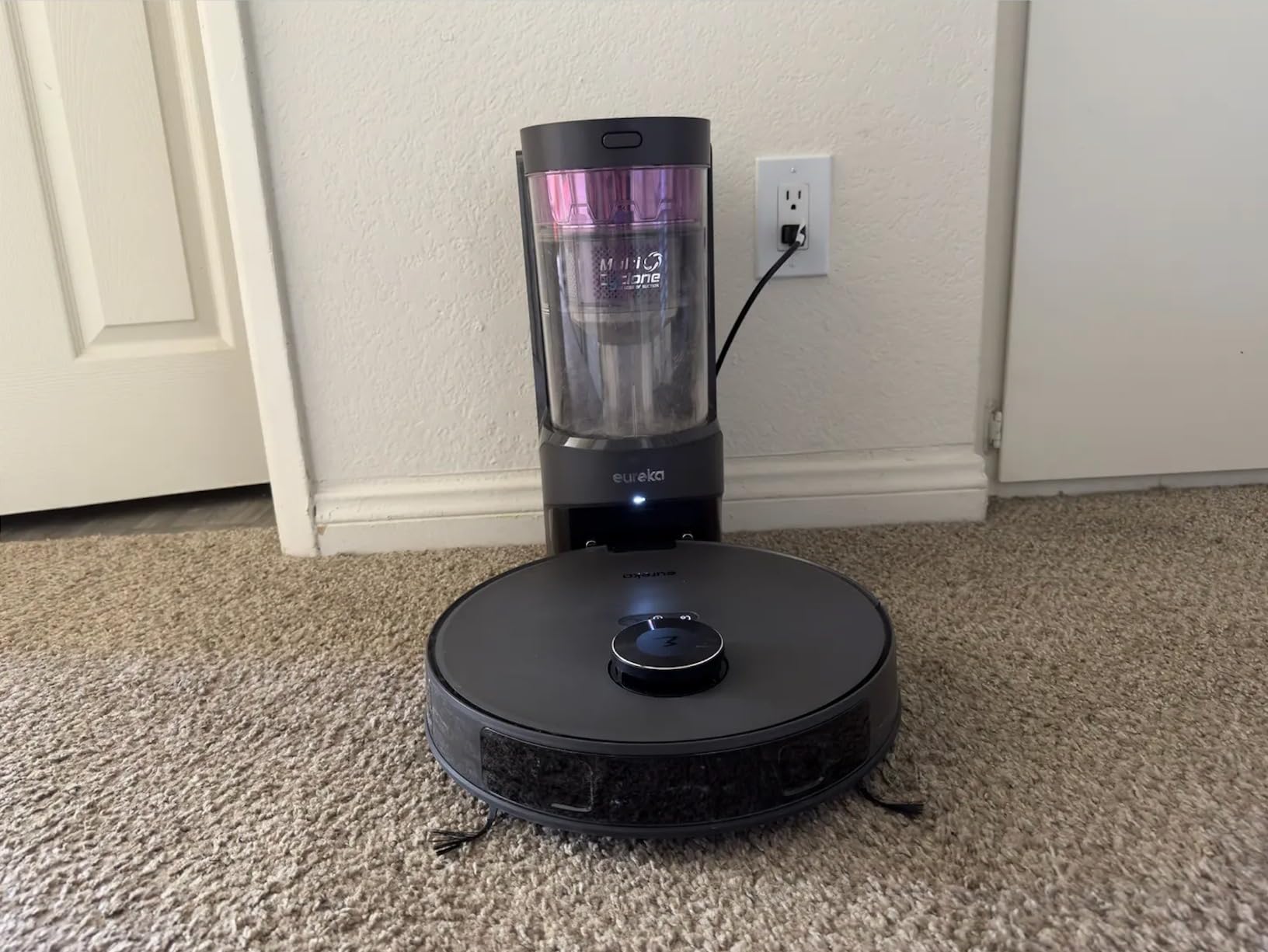 Eureka E10S Review: The robot vacuum is on a table at its docking station with a clear bin